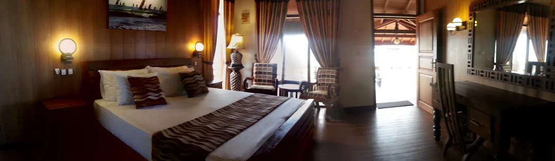 A king room in one sweep: a large bed with wave-patterned cushions, armchairs by the curtained windows, the door open to the veranda and a timber desk and mirror