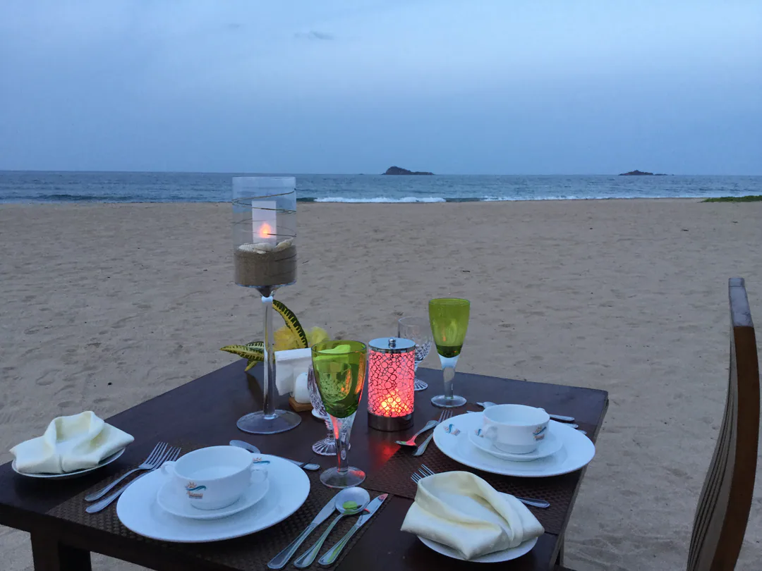A table for two set on the sand at dusk, with candles, coloured glasses and the sea and islands beyond