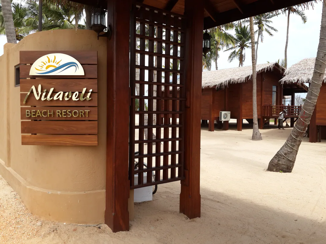 The resort entrance: the Nilaveli Beach Resort sign on a sand-coloured wall beside a timber gate, with cabanas beyond