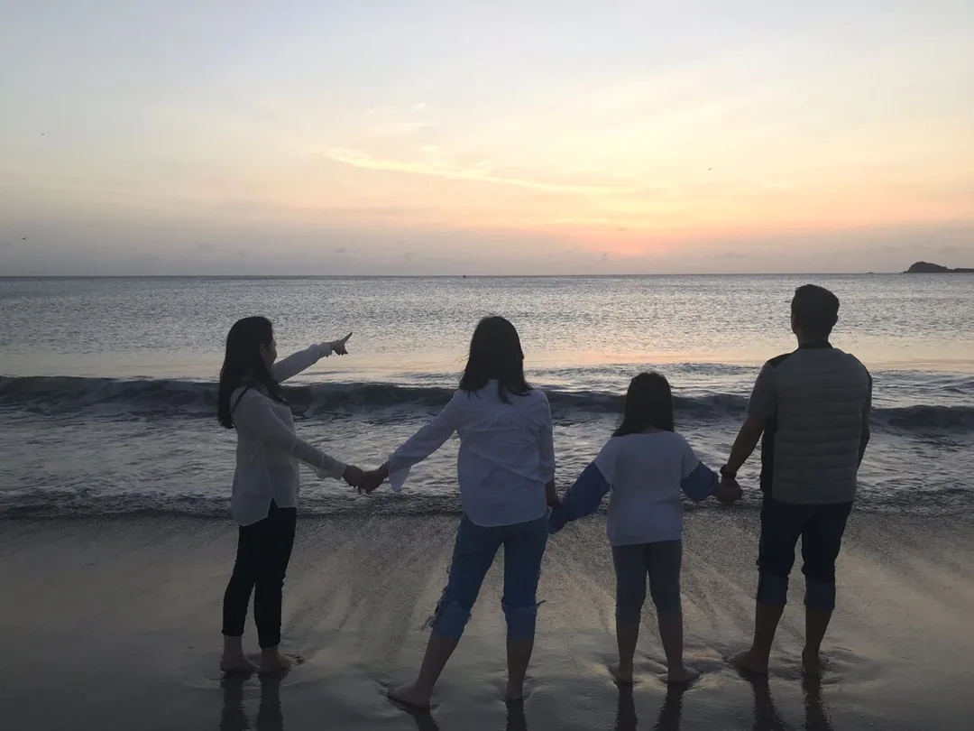 A family holding hands at the water’s edge at sunrise, pointing out to sea