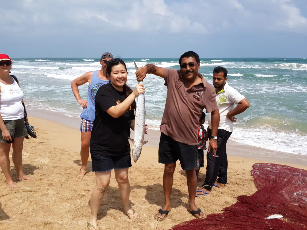 A guest and a fisherman holding up a freshly caught fish on the beach, beside the hauled-in net