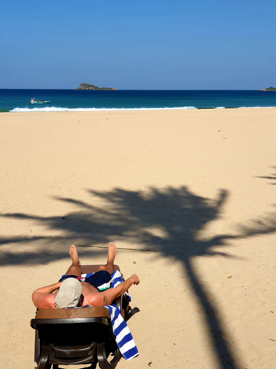 A guest relaxing on a sun lounger in the shade of a palm on the sand, with the sea and Pigeon Island beyond