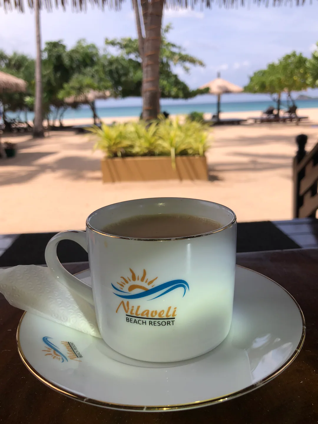 A cup of tea with the Nilaveli Beach Resort logo, looking out to the beach, palms and sea