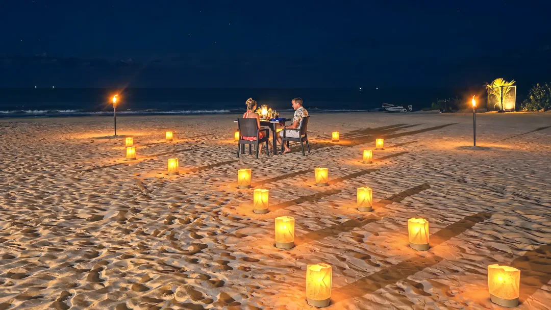 A couple dining at a table on the sand, ringed by lanterns, beside the sea at night