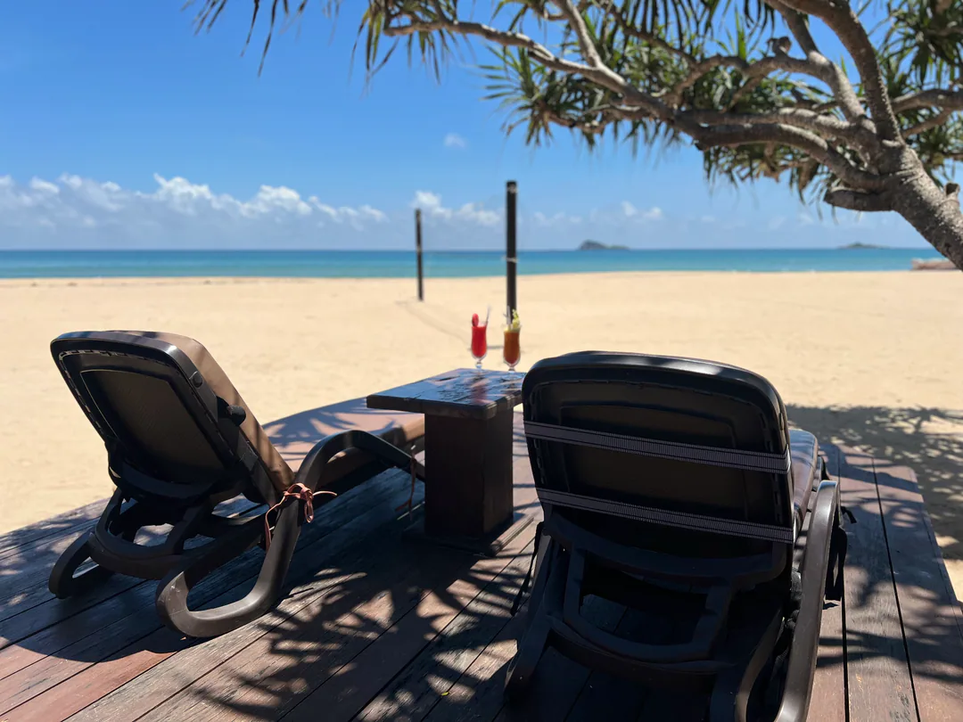 Two cocktails on a small table between loungers on the beach deck, shaded by a pandanus tree
